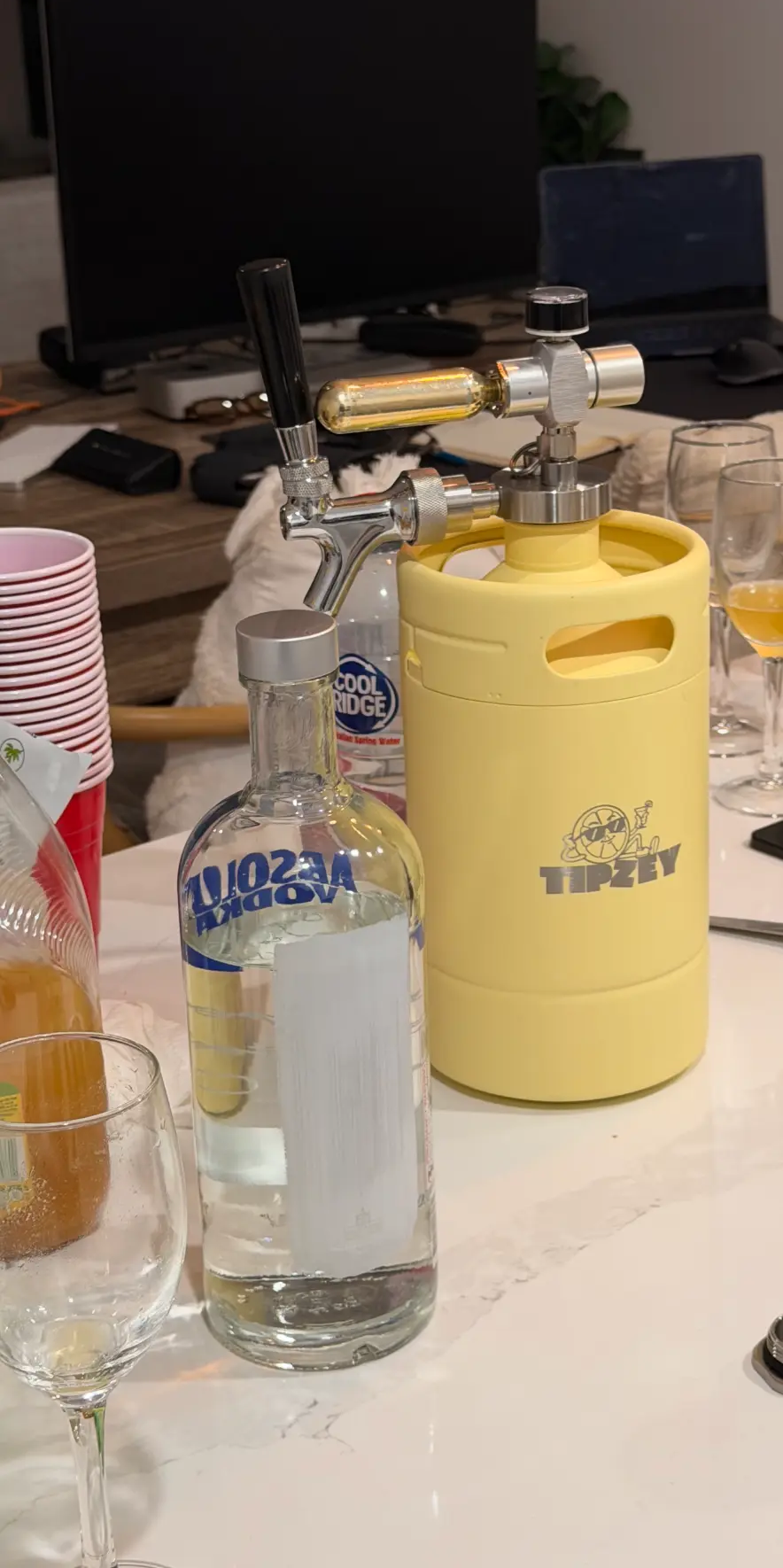 A pale yellow Tipzey mini keg with the tap fitted, on a countertop with drinks and cups